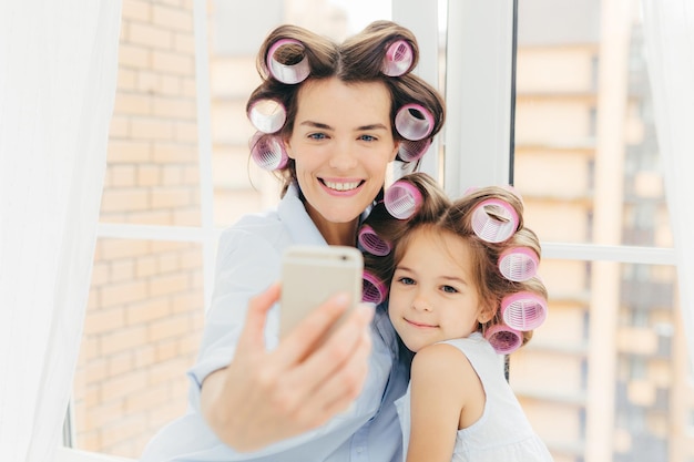 Foto interna da mãe de aparência agradável com expressão positiva e sua filha faz penteados encaracolados posam para selfie no celular moderno se preparam para o carnaval ou festa Mulheres e conceito de beleza