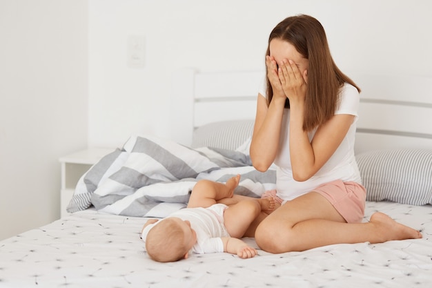 Foto interna da mamãe brincando de esconde-esconde e doente com sua filha pequena enquanto está sentado na cama, mulher com cabelos escuros cobrindo os olhos com as palmas das mãos, vestindo camiseta casual branca.