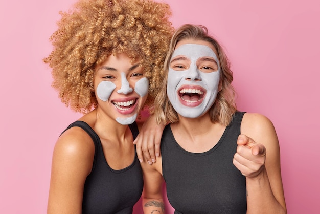 Foto interna de amigas felizes passam por procedimentos de beleza para tratamento de pele aplicar máscaras de argila rir vestida positivamente com camiseta preta casual apontar para a câmera isolada sobre fundo rosa do estúdio