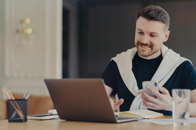 Foto interna de freelancer homem feliz usa gadgets modernos pesquisa informações em laptop hods poses de smartphone modernas na área de trabalho em instalações internas aconchegantes Novo aplicativo usa internet