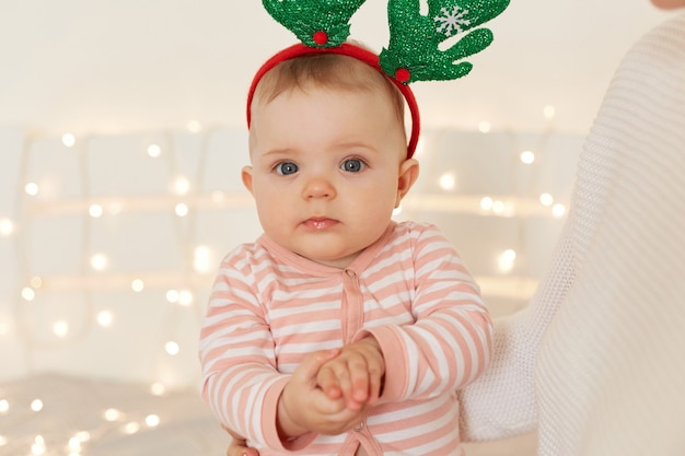 Foto foto interna de lindo adorável bebê infantil vestindo dorminhoco listrado e aros de veado, olhando para a câmera com uma expressão facial encantadora, sentado nos braços da mamãe com luzes de guirlanda no fundo.