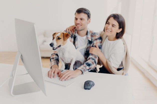 Foto interna de marido e mulher alegres olham com expressões alegres riem enquanto assistem a um filme engraçado descansando juntos no tempo livre cão curioso olha atentamente para o monitor dos teclados do homem do computador