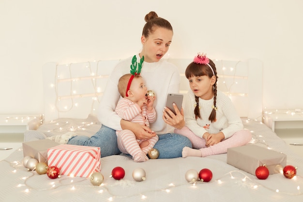 Foto interna de mulher com seus filhos, vestindo trajes casuais, sentada na cama e usando um telefone inteligente, expressando espanto, lendo a mensagem com expressão facial chocada.