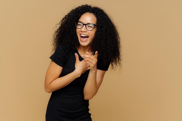 Foto interna de mulher muito feliz ri alto percebe que cenas engraçadas apontam para a câmera mantém a boca aberta usa óculos transparentes camiseta preta casual isolada sobre fundo marrom