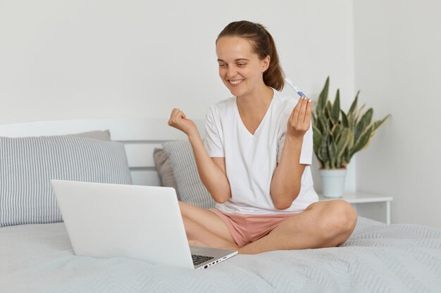 Foto interna de mulher sorridente sentada na cama e conversando por videochamada no laptop contando sobre sua gravidez ostentando resultado olhando para a tela do dispositivo com os punhos cerrados e comemorando boas notícias