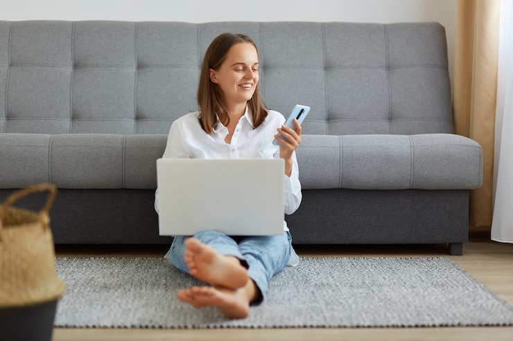 Foto foto interna de mulher sorridente, vestindo camisa branca e jeans, trabalhando online através do laptop em casa, fazendo uma pausa, verificando as redes sociais, e-mail, usando o smartphone, sentado no chão na sala de luz.