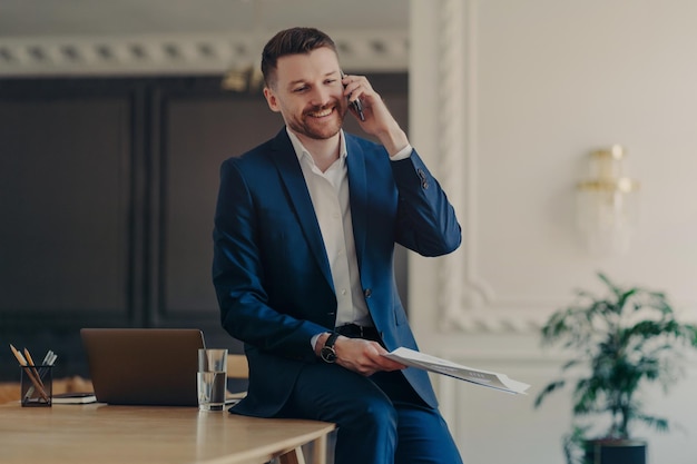 Foto interna de um empresário positivo vestido com um terno formal tem uma conversa por telefone e discute questões de trabalho junto com o parceiro se inclina na mesa segura documentos em papel se prepara para a apresentação