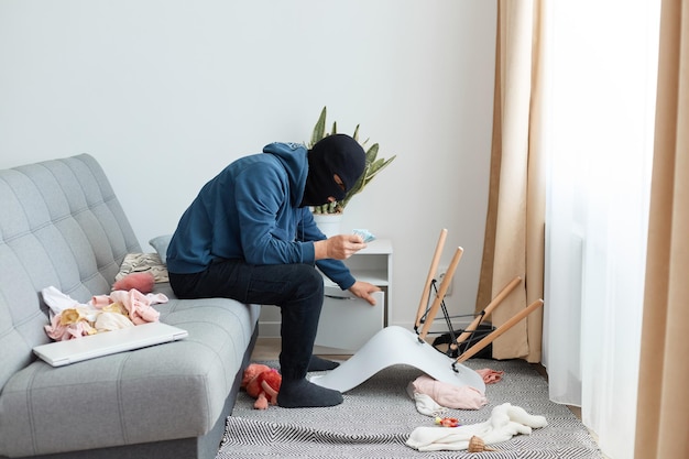 Foto interna de um ladrão masculino usando um capuz azul escuro e máscara preta, um ladrão criminoso roubando a casa de alguém, encontrou o dinheiro escondido na mesa de cabeceira, satisfeito com sua descoberta.