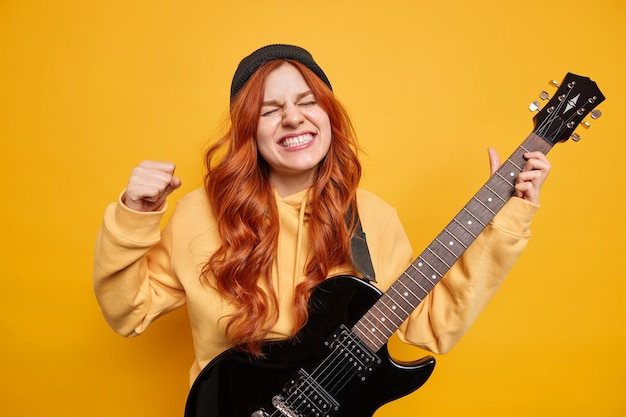 Foto interna de uma adolescente ruiva emocional cerrando os punhos e os dentes usando um capuz com capuz preto e mantendo os olhos fechados sendo uma estrela do rock n roll tocando música na guitarra elétrica preta na parede amarela