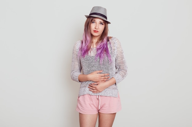 Foto interna de uma jovem atraente com cabelo violeta, usando chapéu, sofrendo de dor de estômago ou dor menstrual, tem problemas de saúde, isolados sobre fundo cinza.