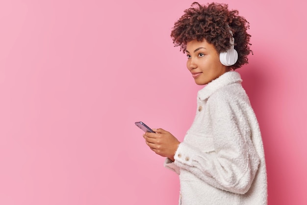 Foto interna de uma jovem de cabelos cacheados que escuta música com fones de ouvido usa tecnologias modernasdres vestida com um casaco de pele branco isolado sobre um fundo rosa com espaço em branco para seu anúncio