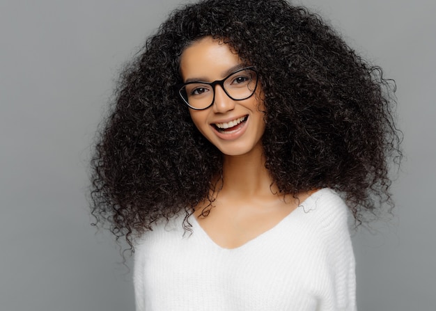 Foto interna de uma jovem feliz com penteado afro, sorrindo amplamente, feliz por ser promovido, usando óculos ópticos e suéter branco