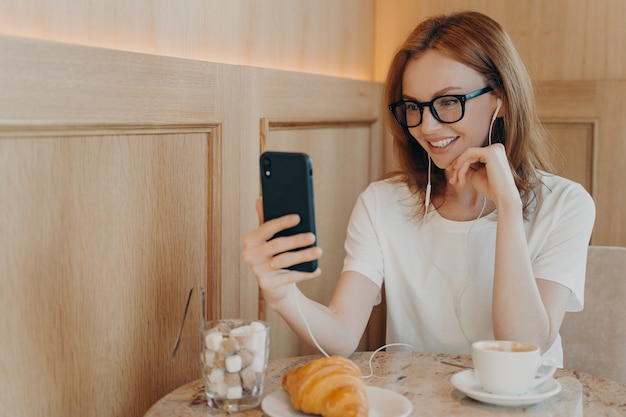 Foto interna de uma jovem ruiva alegre usando telefone celular e fones de ouvido para uma conversa online