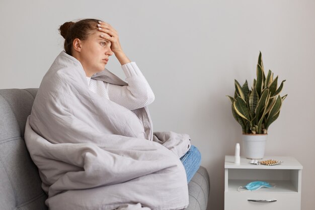 Foto interna de uma mulher insalubre com coque de cabelo sentada no sofá enrolada em um cobertor, tem temperatura alta, mantendo a mão na testa, precisa de tratamento, sofrendo de uma terrível dor de cabeça.