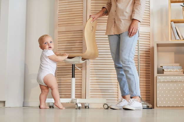 Foto de irreconocible mujer vistiendo ropa casual ayudando a herl niño pequeño a aprender a pararse y caminar