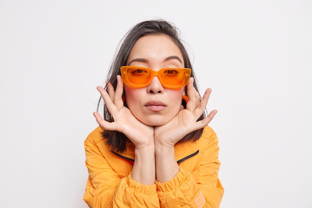 Foto isolada de mulher asiática séria com as mãos nas bochechas, usa óculos escuros laranja tredny e jaqueta usa roupas da última moda, isoladas diretamente sobre a parede branca