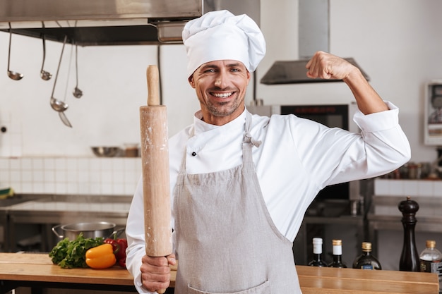 Foto de jefe masculino alegre en uniforme blanco sosteniendo un rodillo, mientras está de pie en la cocina en el restaurante