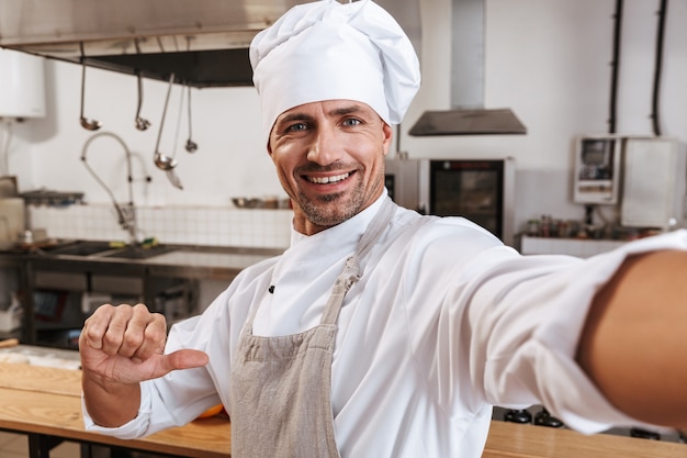 Foto del jefe masculino sonriente en delantal tomando selfie, mientras está de pie en la cocina en el restaurante