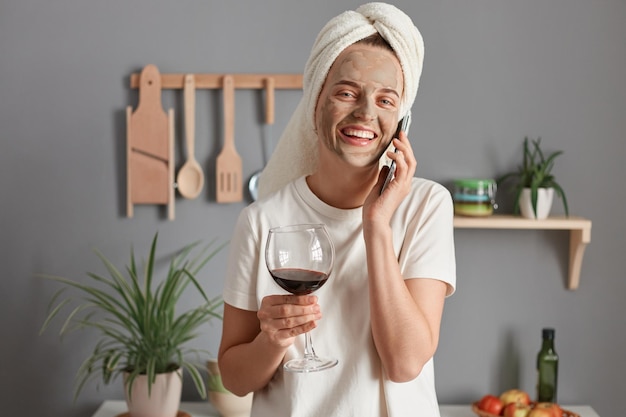 Foto de una joven adulta extremadamente feliz con una máscara cosmética en una toalla blanca envuelta alrededor de la cabeza bebiendo vino en la cocina hablando por teléfono inteligente sonriendo alegremente