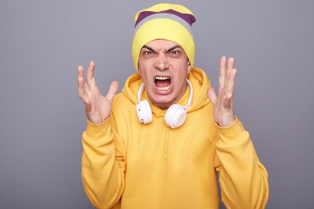 Foto de un joven adulto extremadamente enojado con sombrero de beanie y sudadera con capucha informal amarilla de pie aislado sobre fondo gris gritando con agresión levantó los brazos tiene pelea