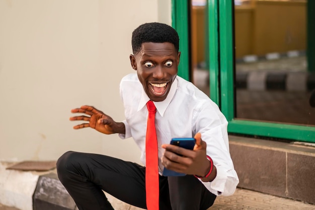 Foto de un joven africano que parece emocionado mientras sostiene un teléfono celular con una camisa blanca y una corbata roja