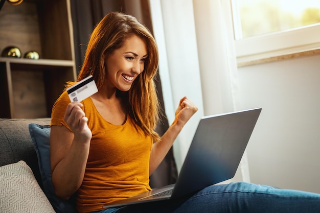 Una foto de una joven alegre que hace compras en línea en su laptop mientras se sienta en el sofá de su casa.