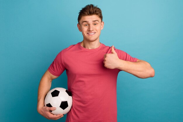 Foto de un joven alegre que sostiene una pelota de fútbol muestra el pulgar hacia arriba perfecto sí anuncia aislado sobre fondo de color azul