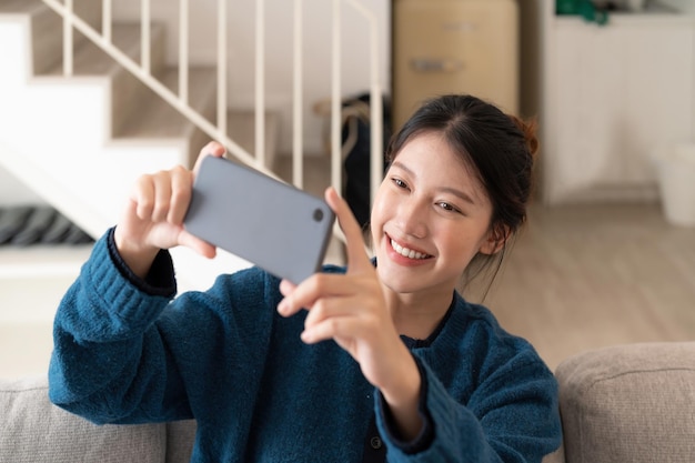 Foto de una joven asiática tomando selfie en un teléfono celular mientras se sienta en un sofá en un apartamento luminoso