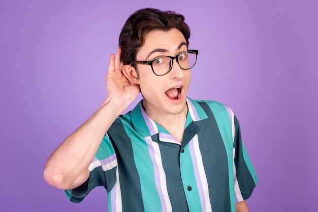 Foto de un joven asombrado con el brazo cerca de las orejas escuchando información chismosa aislada sobre un fondo de color violeta