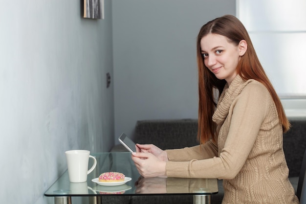 Foto de una joven y atractiva mujer sonriente con suéter sentada en el interior de una mesa en un ambiente agradable