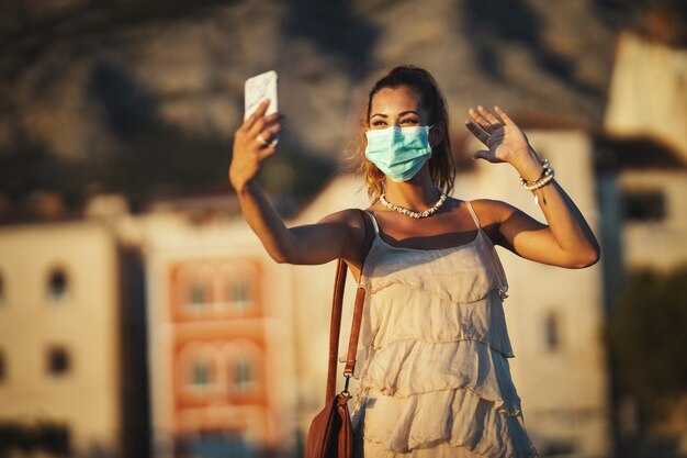 Foto de una joven atractiva que usa una máscara quirúrgica y hace una videollamada con su teléfono inteligente mientras explora la maravillosa ciudad de Meditteranian.