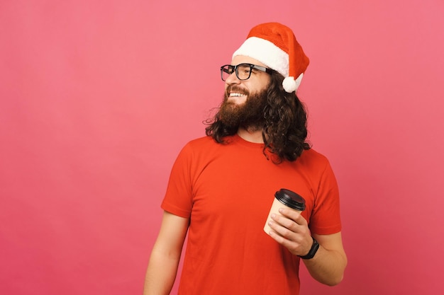 Foto de un joven con barba y cabello largo sosteniendo una taza de café para llevar y mirando el espacio de copias y usando un sombrero de santa claus