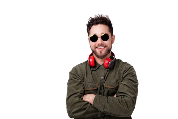 Foto de joven barbudo con gafas de sol y auriculares, de pie con las manos cruzadas sobre la pared blanca