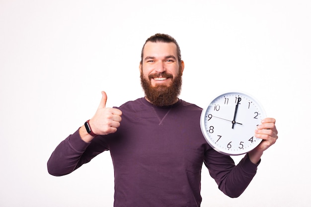 Foto de un joven barbudo sosteniendo un gran reloj blanco y está mostrando un pulgar hacia arriba sonriendo