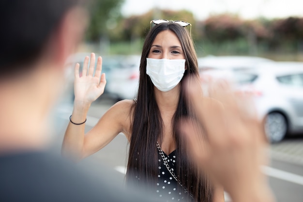 Foto de una joven y bella estudiante con mascarilla quirúrgica y agitando la mano para saludar a su amiga en el campus universitario en el brote de Covid19. Distanciamiento social y nuevo concepto de normalidad.