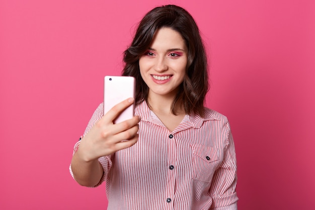 Foto de la joven y bella mujer de pelo oscuro de pie aislado sobre rosa haciendo selfie a través del teléfono móvil, mirando sonriente a la pantalla del dispositivo, viste camisa con rayas. Gente y tecnología concet.