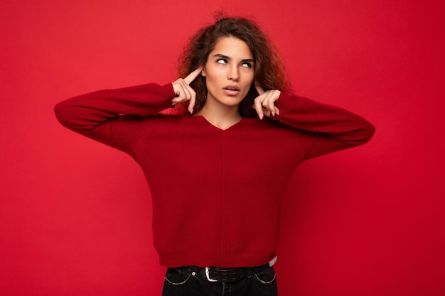 Foto de joven cansada triste insatisfecha atractiva morena mujer de cabello ondulado con emociones sinceras vistiendo suéter rojo aislado sobre fondo rojo con espacio vacío y cubriendo las orejas con los dedos.