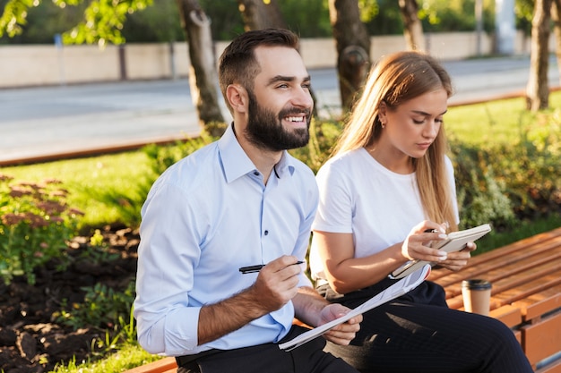Foto de un joven concentrado positivo optimista hombre y mujer empresarios afuera en el parque natural escribiendo notas en cuadernos.