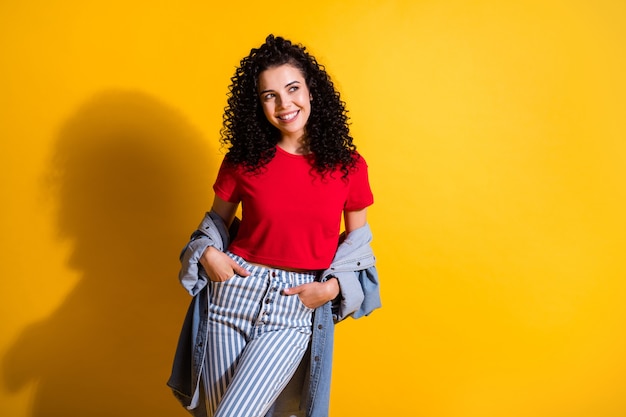 Foto de la joven dama encantadora palmas bolsillos mirar espacio vacío usar chaqueta de jeans a rayas superior rojo aislado fondo de color amarillo