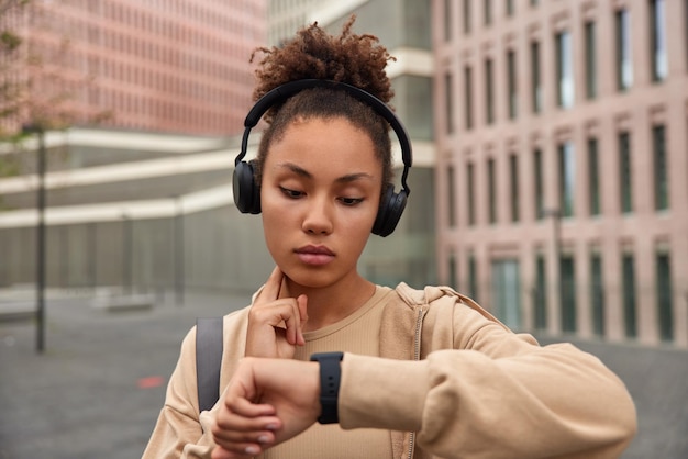 Foto de una joven deportiva con una expresión seria en la cara que toca el cuello y comprueba la frecuencia cardíaca enfocada en un reloj inteligente vestido con una sudadera con capucha escucha música a través de auriculares posa al aire libre contra el edificio de la ciudad