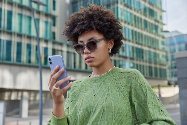 La foto de una joven elegante de cabello rizado usa gafas de sol y cheques de puente verdes recibidos en un mensaje de correo electrónico navega por el sitio web en las redes sociales se encuentra al aire libre contra edificios modernos en la calle urbana