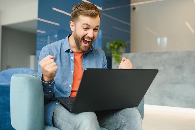 Foto de joven emocionado feliz sonrisa positiva celebrar victoria éxito puños manos sentarse sofá casa trabajo remoto