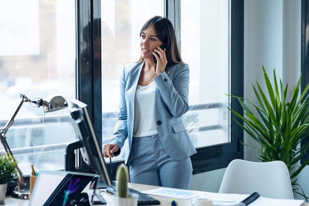 Foto foto de joven empresaria inteligente hablando con su teléfono móvil mientras está de pie mirando por la ventana en el espacio de trabajo moderno.