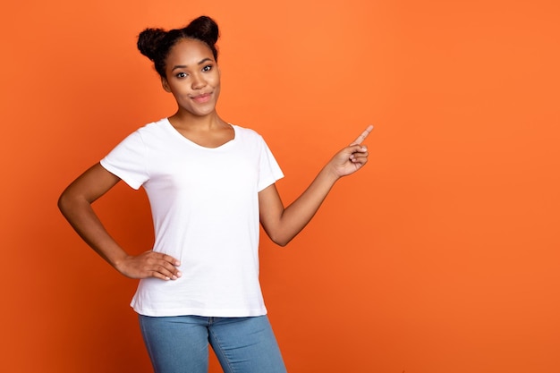 Foto de una joven y encantadora mujer africana que indica un anuncio de decisión de promoción de espacio vacío con el dedo aislado sobre un fondo de color naranja