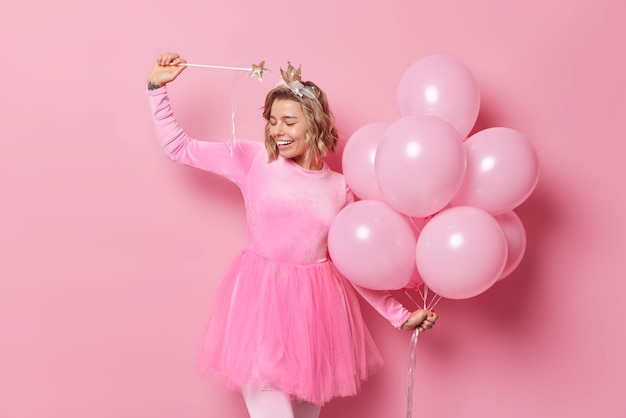 La foto de una joven europea feliz se divierte en poses de fiesta con un montón de globos de helio usa una varita mágica para hacer que tus sueños se hagan realidad poses contra un fondo rosa. Concepto de personas y vacaciones