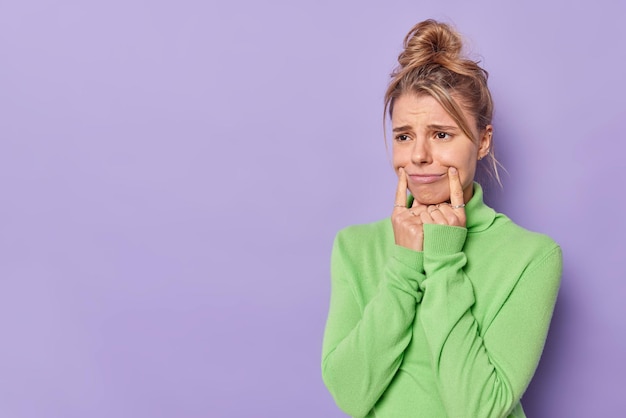 La foto de una joven europea molesta estira la boca en una sonrisa forzada tiene una expresión triste y usa poses casuales de cuello alto verde sobre un fondo púrpura con espacio de copia para su contenido publicitario