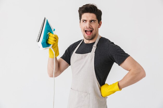 Foto de un joven europeo con guantes de goma amarillos para la protección de las manos sonriendo y sosteniendo el hierro mientras limpia la casa aislada sobre blanco