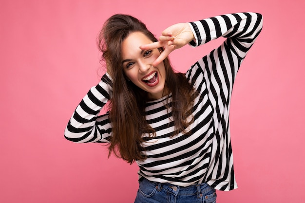 Foto de joven europeo positivo sonriente encantadora alegre feliz hermosa mujer morena con emociones sinceras vistiendo suéter de rayas casual aislado sobre fondo rosa con espacio de copia.