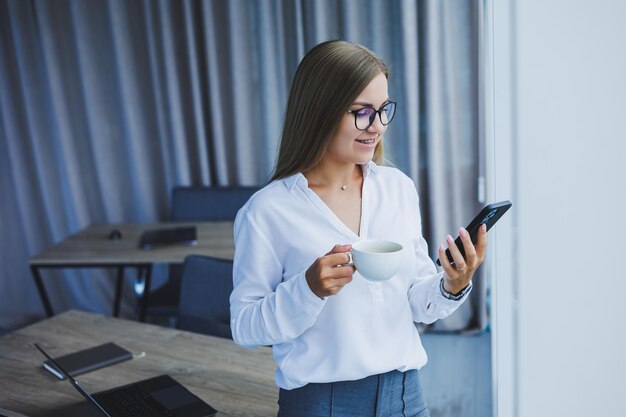 Foto de una joven exitosa con una camisa sonriendo y trabajando en una computadora portátil mientras habla por teléfono en una oficina moderna con ventanas grandes Trabajo remoto con café