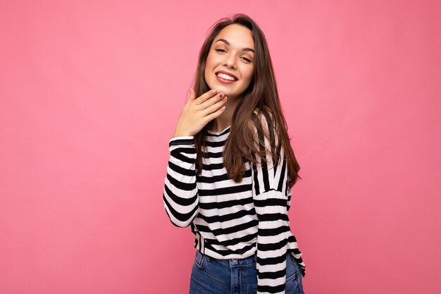 Foto de joven feliz sonriente positiva hermosa mujer morena con emociones sinceras vistiendo suéter de rayas casual aislado sobre fondo rosa con espacio de copia.